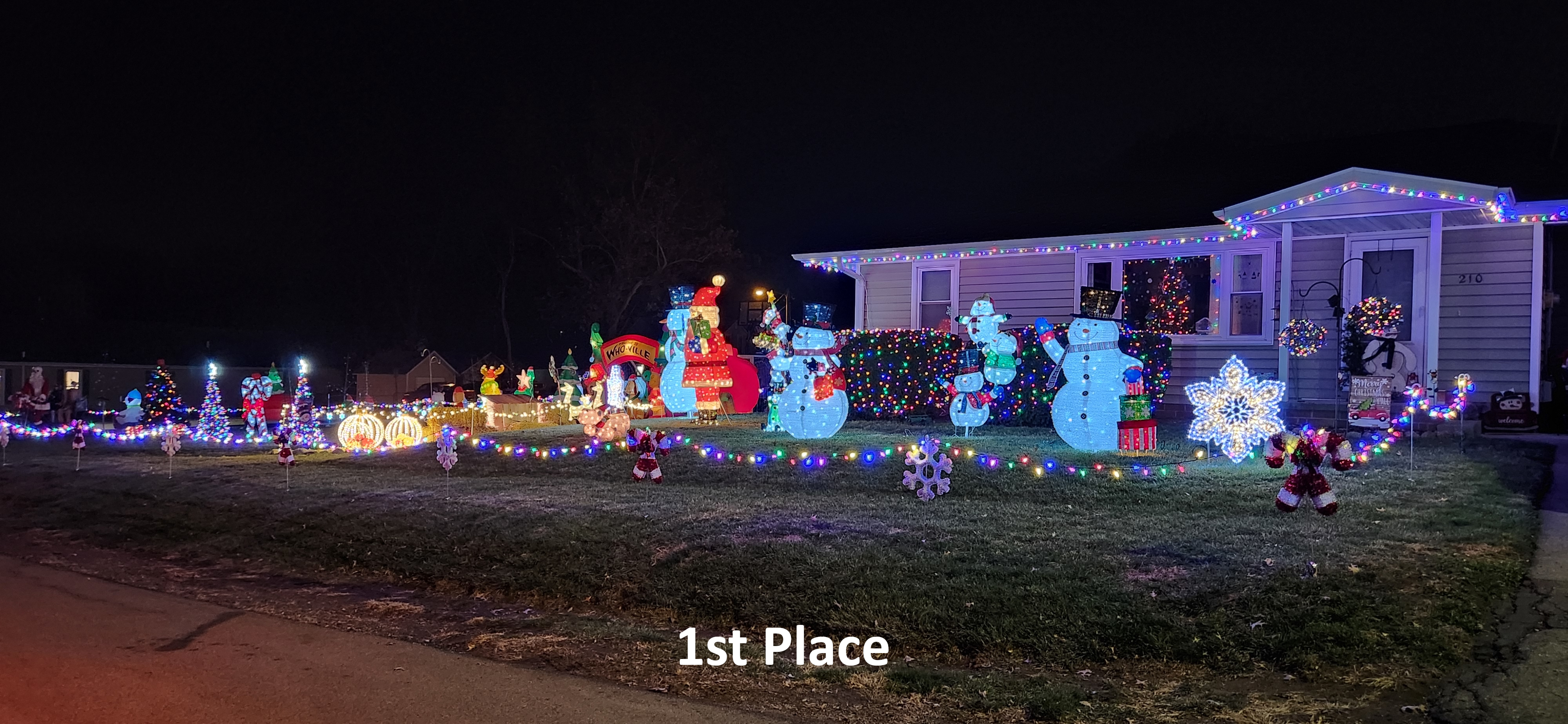 A festive house decorated with colorful Christmas lights as part of the Light Up Carlisle event features bright snowmen, a large Santa, and other seasonal figures. The lawn and roof are adorned with glowing decorations. A sign reads "1st Place.