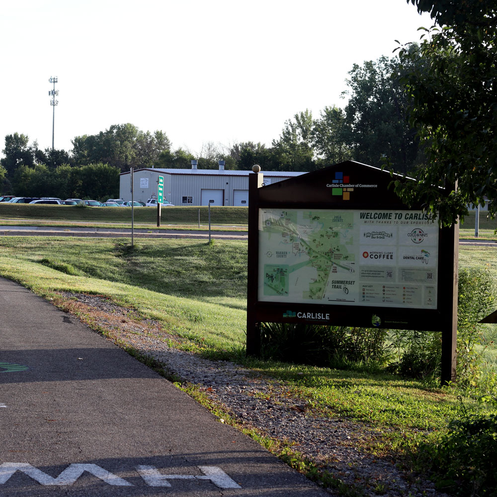 A welcome sign for Carlisle is positioned next to a paved walking path, surrounded by green grass and trees. A building is visible in the background, along with a parking lot. Several informational placards are displayed on the sign.