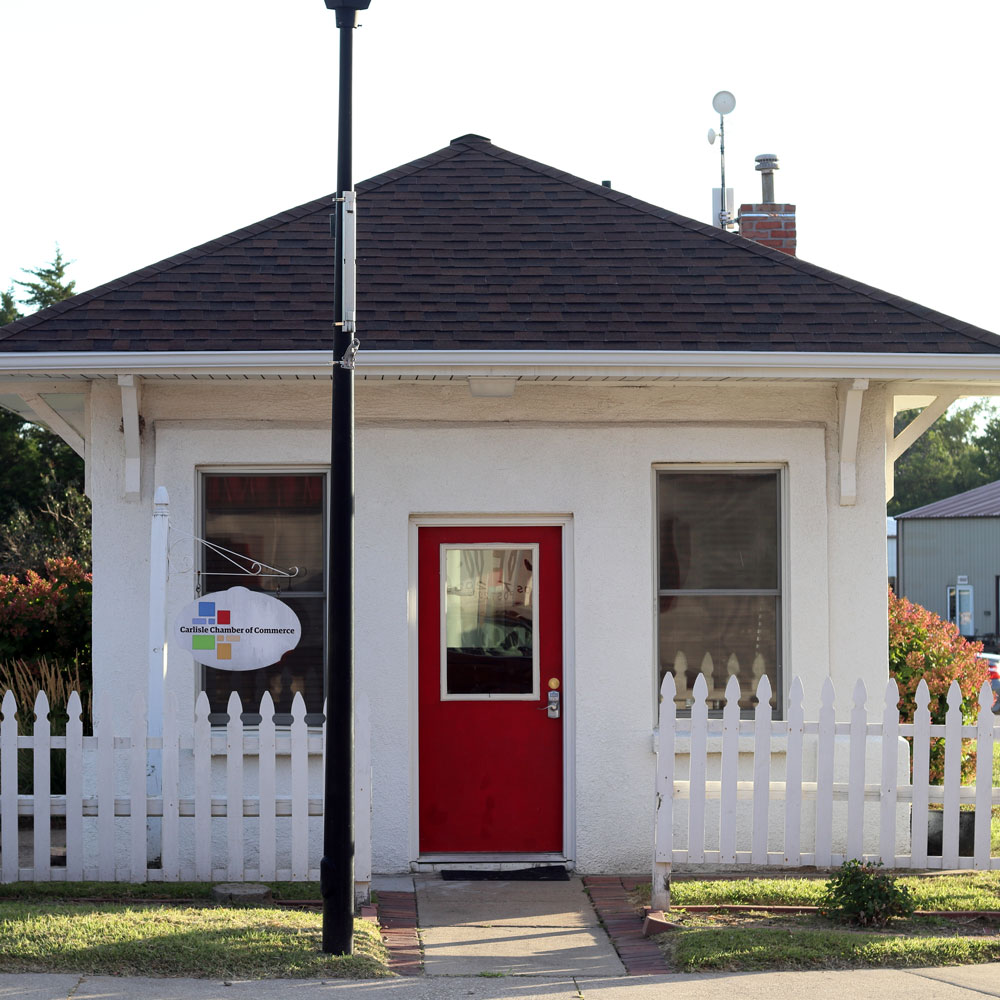 A small white building with a dark brown roof stands behind a white picket fence. It has two large windows flanking a red door with a sign above it. A lamp post is located in front of the building. The sign near the door reads "Chamber of Commerce.