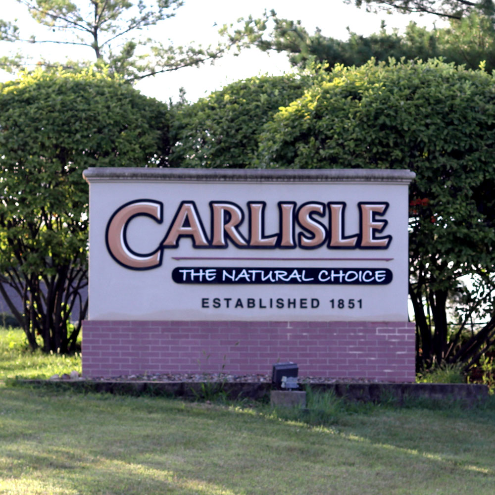 A large outdoor sign reads "Carlisle, The Natural Choice, Established 1851" against a backdrop of shrubs and trees. The sign has a brick base and a beige upper part with bold, stylized text.