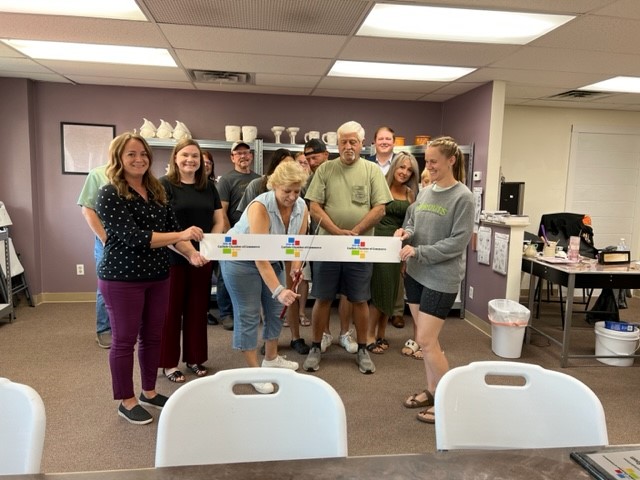 A group of people smile and gather in a room for a ribbon-cutting ceremony. Two women in front hold a ribbon. The room has a lavender wall and shelves with various items in the background. Tables and chairs are visible in the foreground.