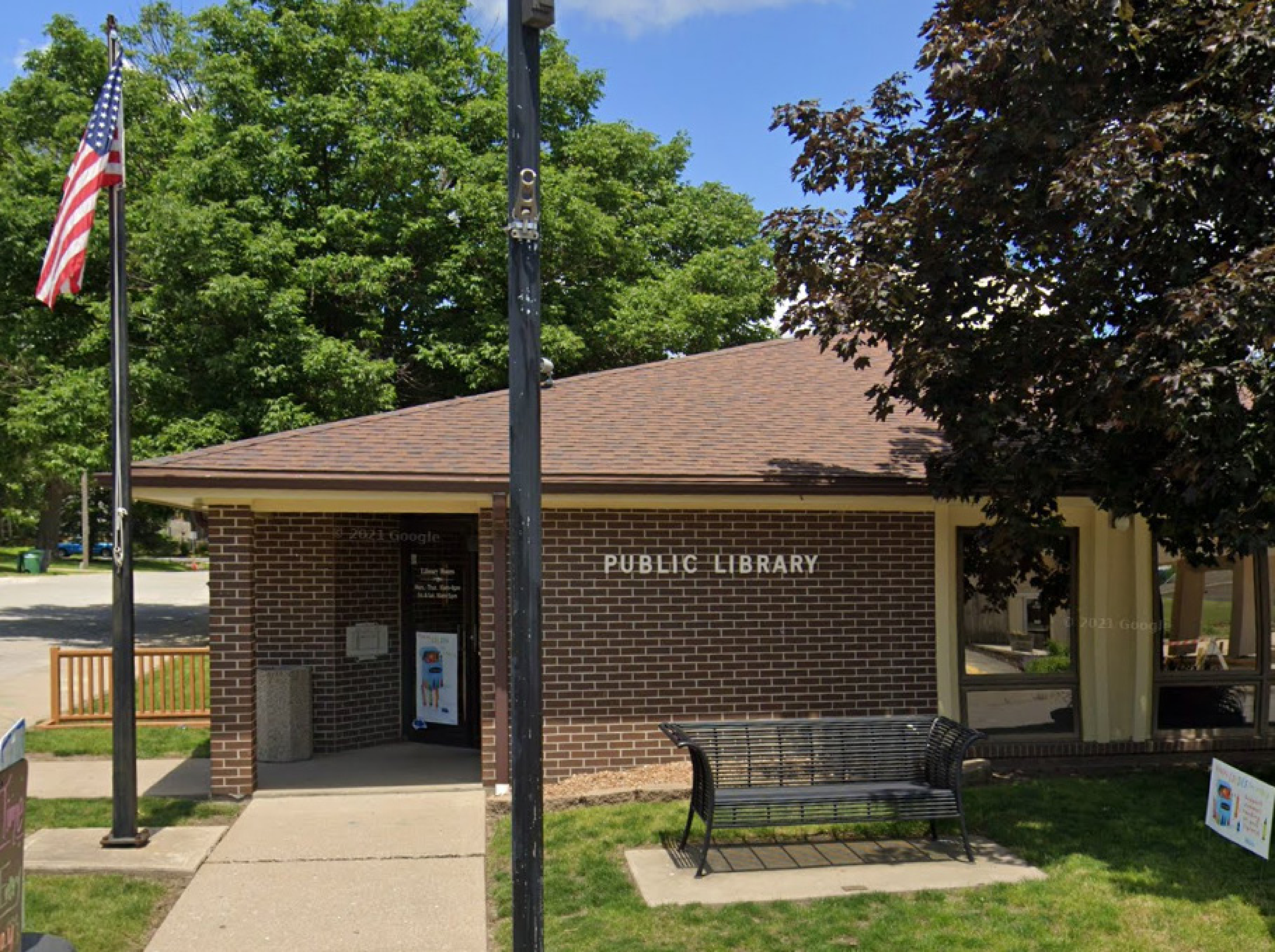 A small public library building with a brick exterior, a sloped roof, and an American flag on a pole nearby. There is a bench on the grass in front of the entrance, and trees surrounding the area. The entrance has a glass door with signage visible.