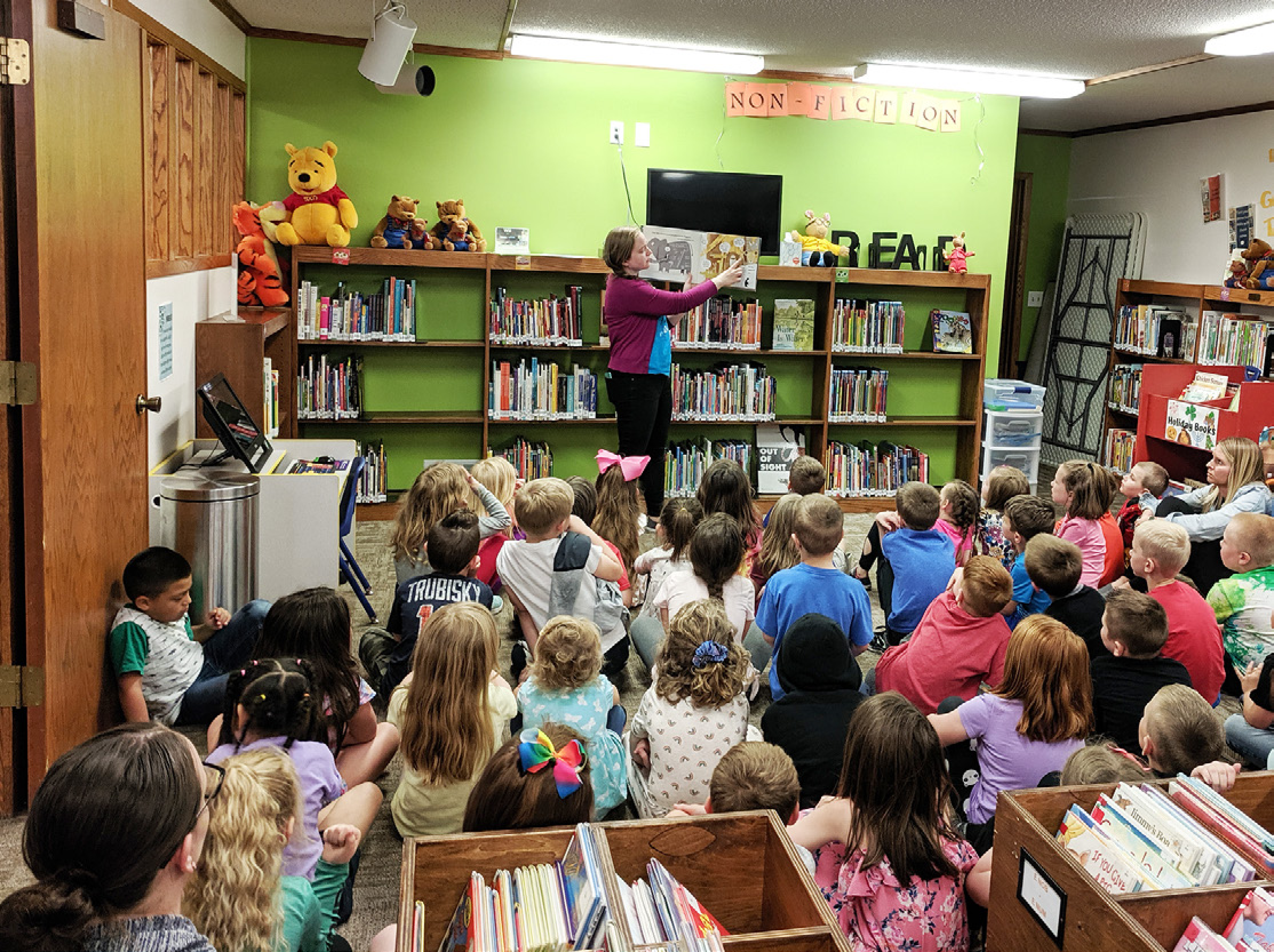 A teacher stands in front of a classroom filled with young children sitting on the floor, attentively listening to a story. The room is colorful with a bookshelf, books, and children's artwork on the walls. The teacher holds a picture book while reading aloud.