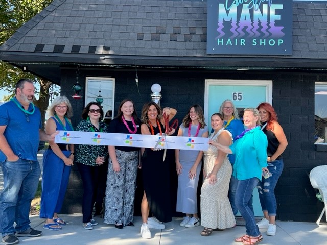 A group of people stands in front of a black building with a sign that reads "Love the Mane Hair Shop." They are holding a long ribbon with scissors prepared for a ribbon-cutting ceremony. The front of the building features a "65" address sign.