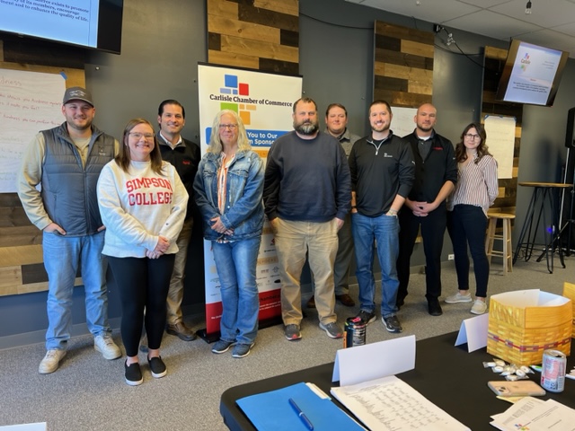 A group of nine people standing indoors in front of a Carlisle Chamber of Commerce banner. The group appears relaxed and is dressed in casual and semi-casual attire. There are tables with papers and a can in the foreground.