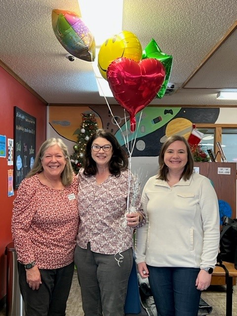 Three women standing indoors, smiling and holding balloons. The balloons are colorful, including a heart-shaped one. The room has festive decorations such as a small Christmas tree, and there's a chalkboard with writing in the background.