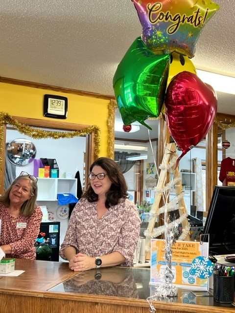 Two women stand behind a counter, smiling. Balloons with "Congrats!" are tied to the counter, alongside decorative Christmas items. A clock on the wall reads 11:26. The counter has various papers and small decorations.