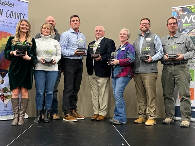 A group of seven people stands on a stage holding awards. They are dressed in business casual attire and smile at the camera. Two banner stands are visible in the background, promoting local tourism and economic development.