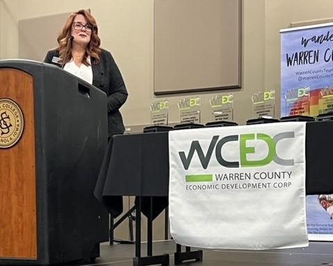 A woman is standing at a podium giving a speech. The podium has a college logo, and several awards are displayed on a table beside her, covered with a cloth that reads "Warren County Economic Development Corp" and the WCDC logo.