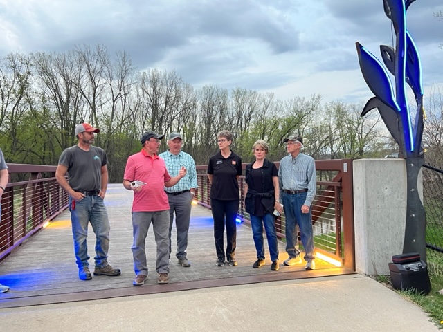 Six people stand and talk on a pedestrian bridge at dusk, near a tall metal sculpture with blue lights. Trees with sparse leaves and a cloudy sky are visible in the background.