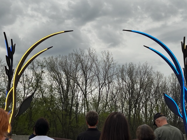 A group of people stand outdoors under a cloudy sky, facing a display of tall, curved sculptures with blue and yellow lights, surrounded by leafless trees in the background.