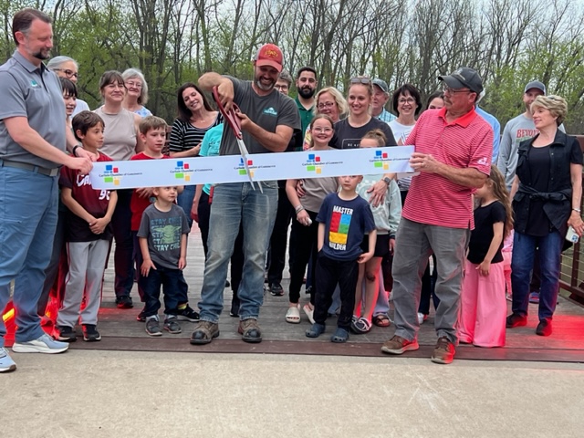 A group of people, including children and adults, smile as a man in a red cap cuts a ribbon with oversized scissors during an outdoor ribbon-cutting ceremony on a wooden bridge. Trees are visible in the background.