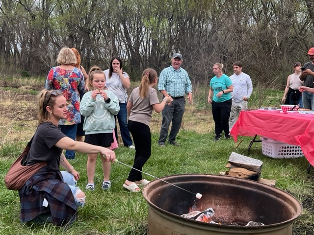 A group of people gather outdoors near a fire pit, roasting marshmallows. Some stand and chat while others sit or toast marshmallows. A table with food is set up nearby. Trees and grass are in the background.