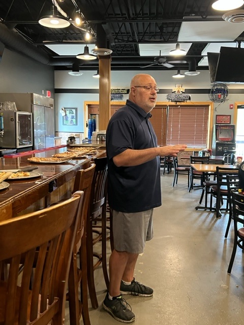 A man wearing glasses, a navy polo shirt, and gray shorts stands next to a bar counter with pizza trays in a casual restaurant, speaking to someone out of frame. Tables and chairs fill the room.
