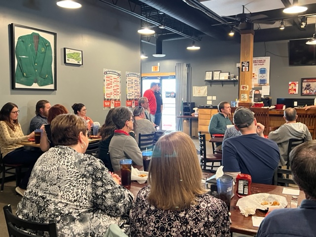 A group of people sits at tables in a casual restaurant, listening to a speaker at the front. Food and drinks are on the tables. A framed green jacket and posters decorate the walls. Light streams in from the open door.