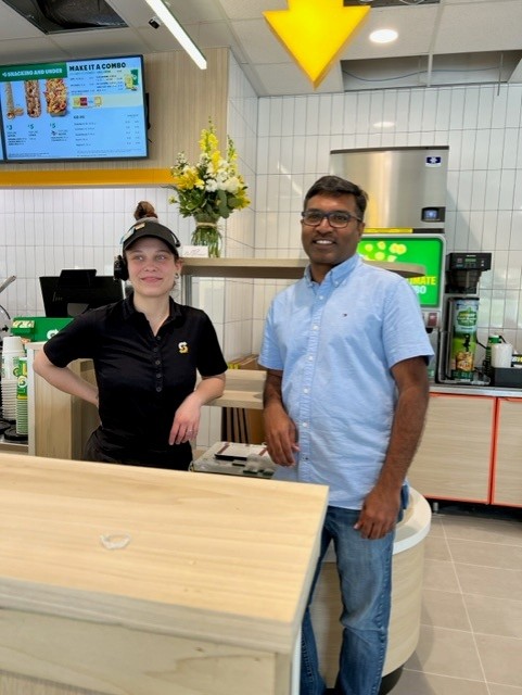 A woman in a black uniform and cap stands behind a counter next to a man in a light blue shirt at a sandwich shop, with a menu board and flower arrangement visible in the background.