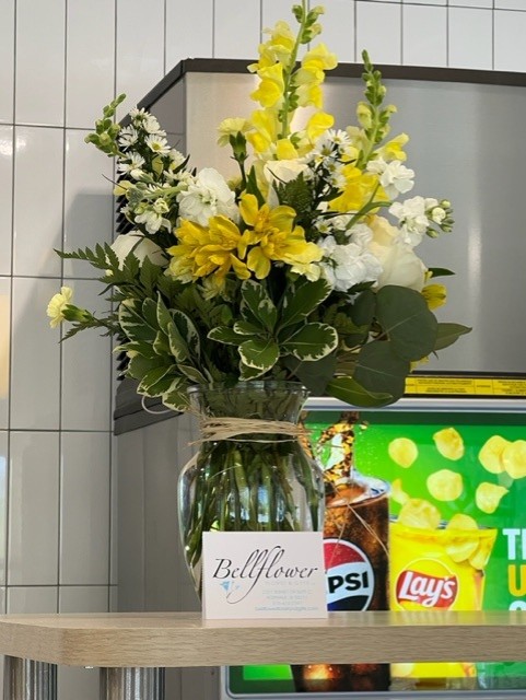 A glass vase filled with yellow and white flowers sits on a counter. Behind it, there's a Pepsi and Lay's vending machine. A card with "Bellflower" written on it is placed in front of the vase.