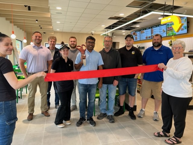 A group of people smiling and posing inside a restaurant for a ribbon-cutting ceremony. One person in the center holds large scissors, and two people at the ends hold the red ribbon. Menu boards are visible in the background.