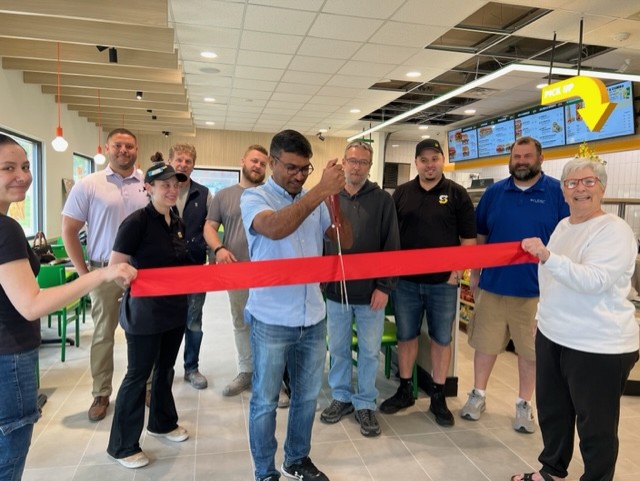 A group of people stand inside a restaurant, smiling as a man in the center uses scissors to cut a red ribbon, celebrating a grand opening. Others hold the ribbon on both sides.