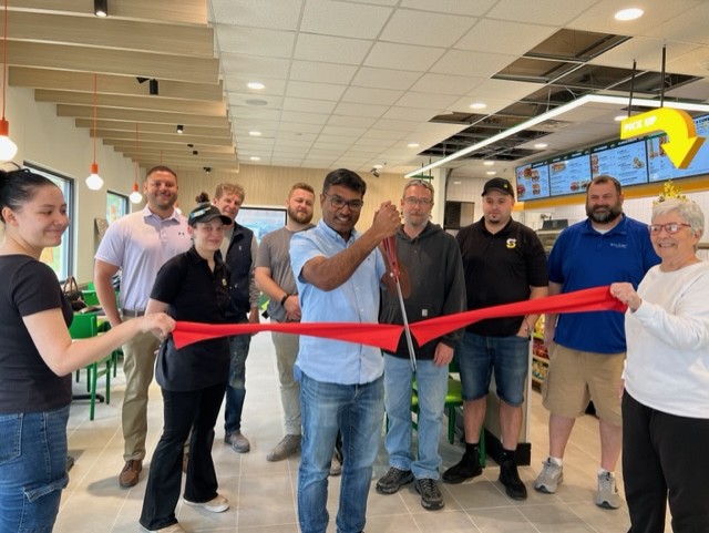 A group of people stands indoors smiling as a man in the center cuts a red ribbon with large scissors, celebrating a store opening. Menu boards and seating are visible in the background.