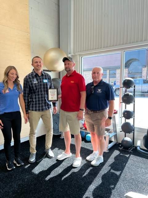 Four adults stand indoors by gym equipment, smiling. One man, holding a framed certificate, stands between two men and a woman. Sunlight shines through large windows behind them, and weights are visible by the wall.