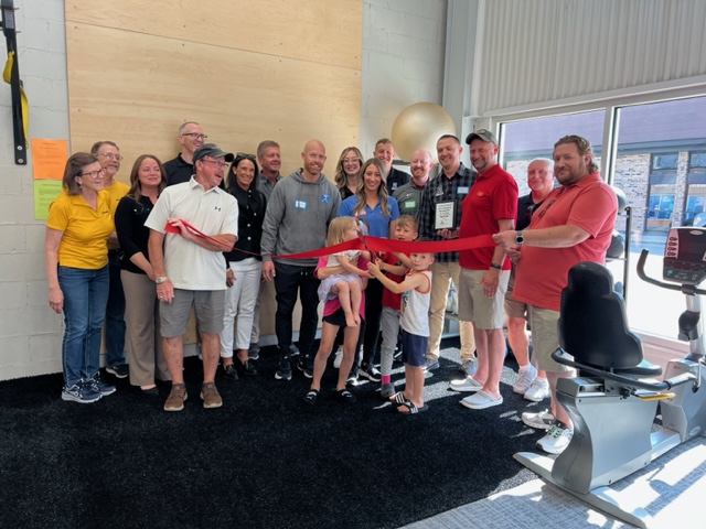 A group of adults and children stand together indoors as two children cut a red ribbon for a celebration or opening event. People are smiling, and exercise equipment is visible on the right.