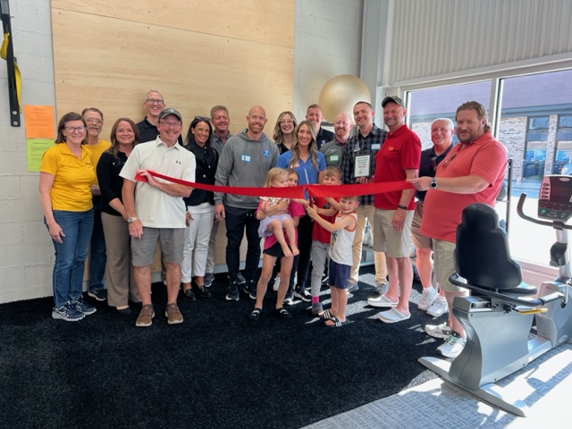 A group of adults and children gather indoors, smiling, as several children cut a red ribbon for a ceremony. Exercise equipment is visible on the right, and sunlight streams in through large windows.