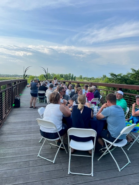 A group of people sits at long tables on a wooden bridge outdoors, enjoying a meal together on a sunny day with a scenic view of greenery and a partly cloudy sky.