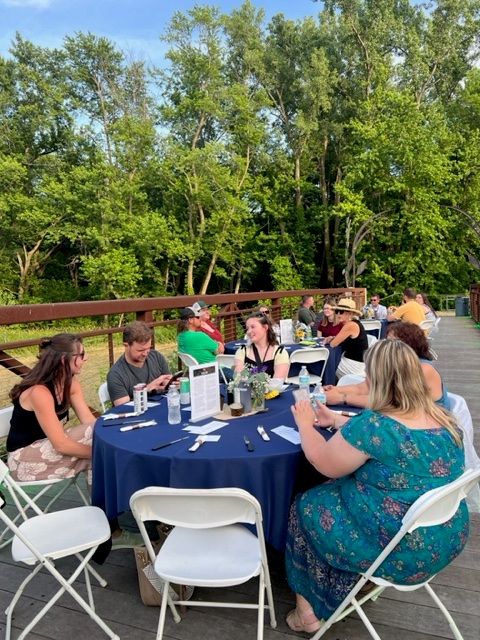 People sit around round tables with blue tablecloths, chatting and using their phones at an outdoor event on a wooden deck, surrounded by green trees on a sunny day.