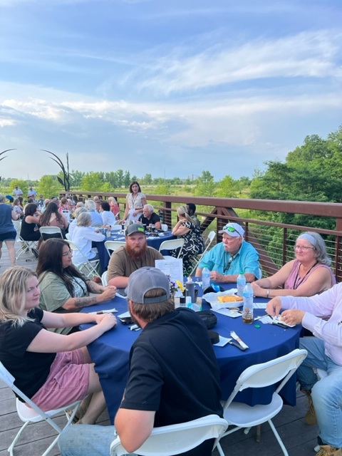 A group of people sit at round tables covered with blue tablecloths, socializing and eating outdoors on a wooden deck with a scenic view of trees and a partly cloudy sky.
