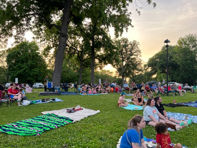 People sit on blankets and lawn chairs in a park at sunset, surrounded by trees and greenery, as they gather for an outdoor event. Some are talking and relaxing, while others look toward the front.