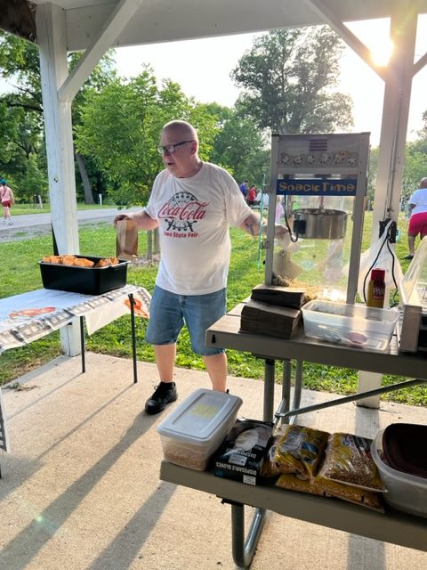 A man in a Coca-Cola t-shirt stands beside a popcorn machine and a table with snacks under a pavilion at a park. The sun shines through the trees in the background.