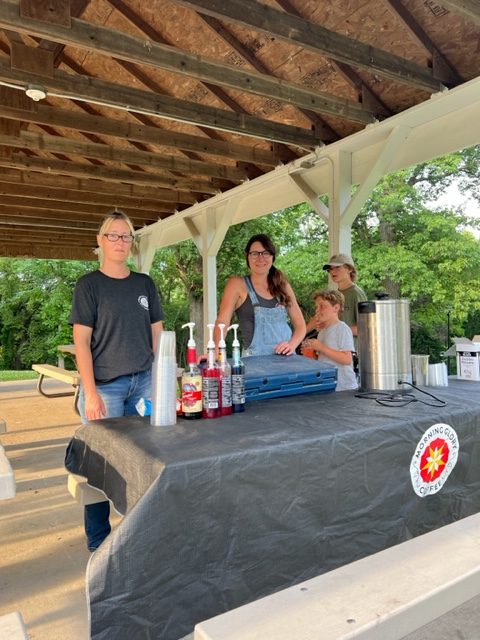 Four people stand behind a table covered with a black cloth displaying colorful syrup bottles, cups, and a metal drink dispenser, under a pavilion in a park on a sunny day.