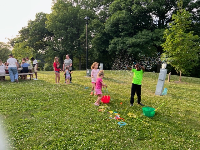 Children play with bubbles on a grassy field while adults and other kids gather near picnic tables in the background. Trees provide shade and bubble wands and buckets are scattered on the ground.
