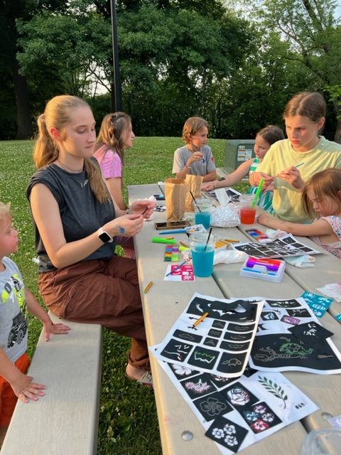 A group of children and teens sit at a picnic table in a park, doing arts and crafts with stencils, paint, and markers. Art supplies and finished artwork are spread across the table. Trees and greenery are in the background.