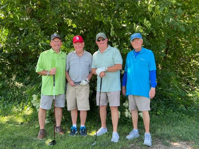 Four older men stand together outdoors in front of green bushes, dressed in golf attire with hats and shorts, each holding or standing with a golf club, smiling at the camera on a sunny day.