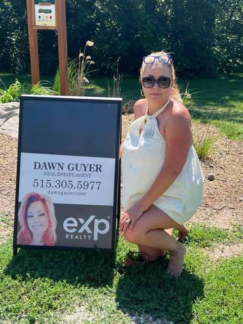 A woman in sunglasses and a white dress kneels next to a real estate sign for Dawn Guyer, exp Realty, outdoors on a sunny day with grass, gravel, and trees in the background.