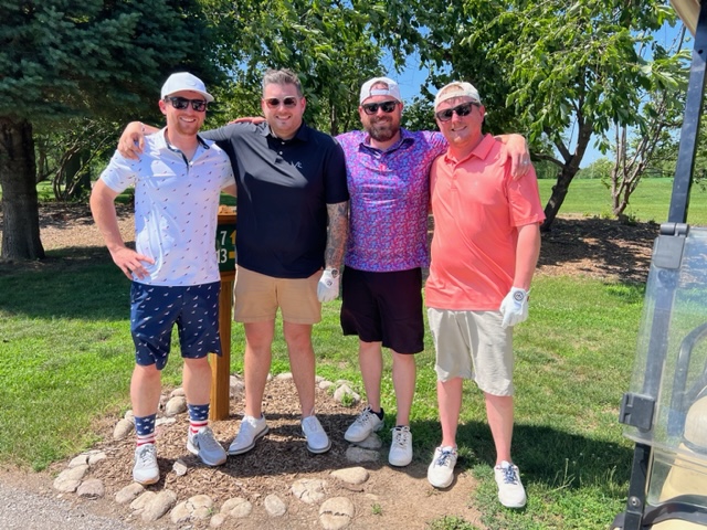 Four men stand outdoors on a golf course, smiling for the camera. They are dressed in casual golf attire with hats and sunglasses, surrounded by grass, trees, and blue sky, with a golf cart partly visible on the right.