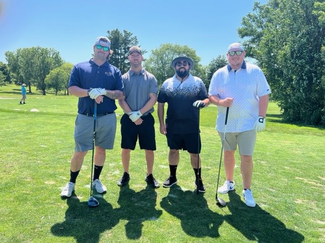 Four men pose together on a sunny golf course, each holding a golf club and wearing golf attire. Trees and green grass surround them, with a clear blue sky overhead.