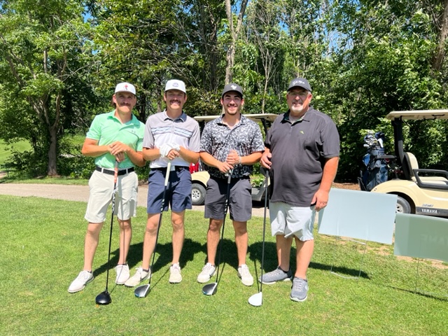 Four men dressed in golf attire stand together on a sunny golf course, smiling and holding golf clubs. Golf carts and trees are visible in the background.
