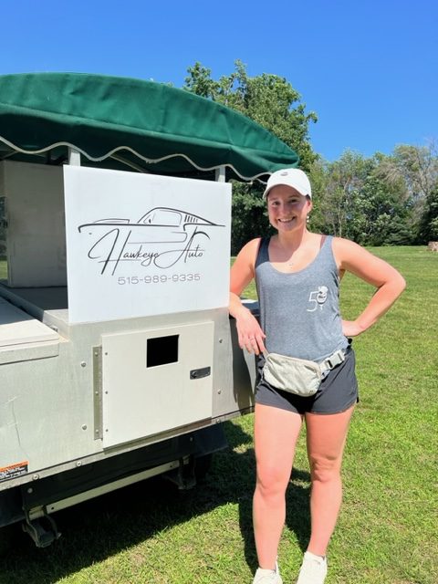 A smiling woman in athletic clothes and a white cap stands next to a white food cart with a green canopy. The cart has a logo reading "Hawkeye Huto" and a phone number. Trees and blue sky are in the background.