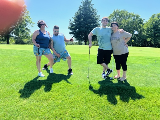 Four people pose together on a sunny golf course, holding golf clubs. Two people stand close together on the right, while the other two pose playfully on the left. A large tree and golf flag are visible in the background.