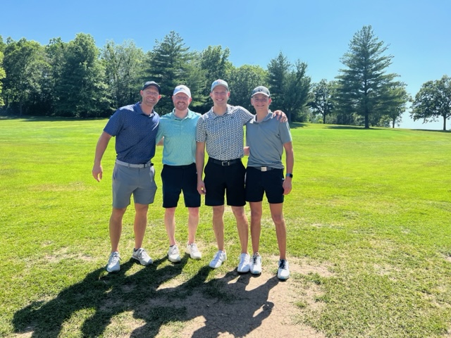 Four men stand together smiling on a sunny golf course, wearing casual golf attire and caps, with green grass and tall trees in the background.