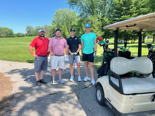 Four men stand on a golf course path next to a golf cart, dressed in casual golf attire, smiling on a sunny day with green grass and trees in the background.