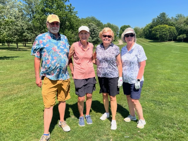 Four older adults stand together smiling on a sunny golf course, dressed in casual summer clothing, with green grass and trees in the background.