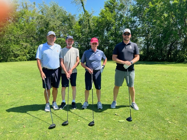 Four men stand on a sunny golf course holding golf clubs, with green grass and trees in the background. They are dressed in casual golf attire and smiling at the camera.