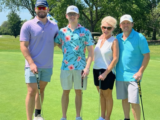 Four people stand on a sunny golf course holding golf clubs, smiling at the camera. They are dressed in colorful, casual summer outfits. Trees, grass, and a golf cart are visible in the background.