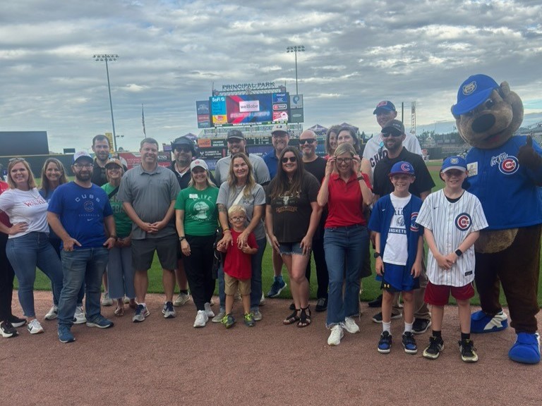 A group of people and a bear mascot pose together on a baseball field, smiling in front of the scoreboard at a stadium. Some are wearing Cubs shirts and hats; children and adults are included in the group.