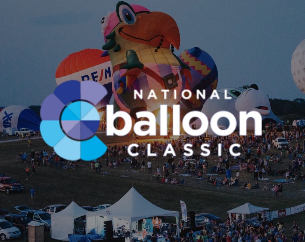 A crowd gathers at sunset to watch colorful hot air balloons, including a large frog-shaped balloon, at the National Balloon Classic festival. Tents and festival booths are visible in the foreground.