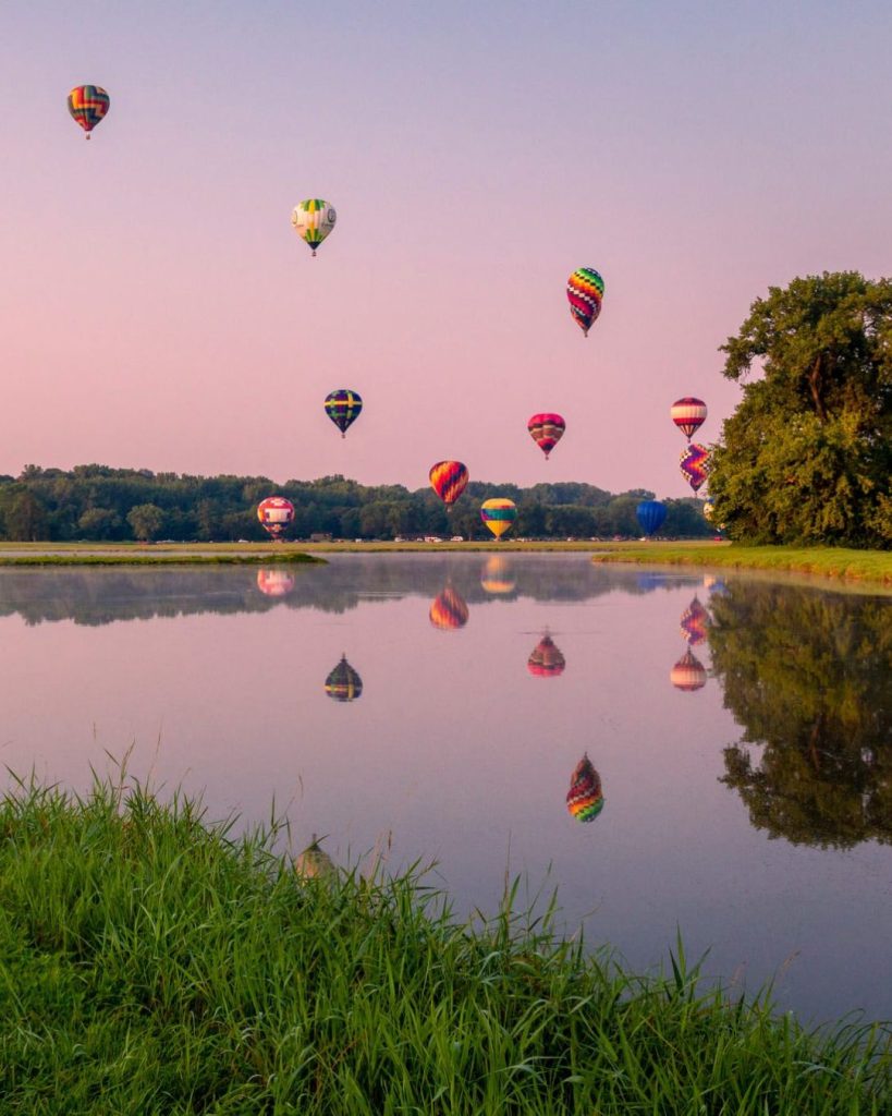 Colorful hot air balloons float above a calm lake at sunrise or sunset, with their reflections visible in the water. Lush green grass frames the foreground and trees line the far shore.